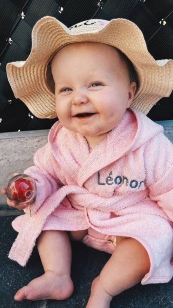 Lachende baby in roze badjas met strohoed en rammelaar zittend op handdoek