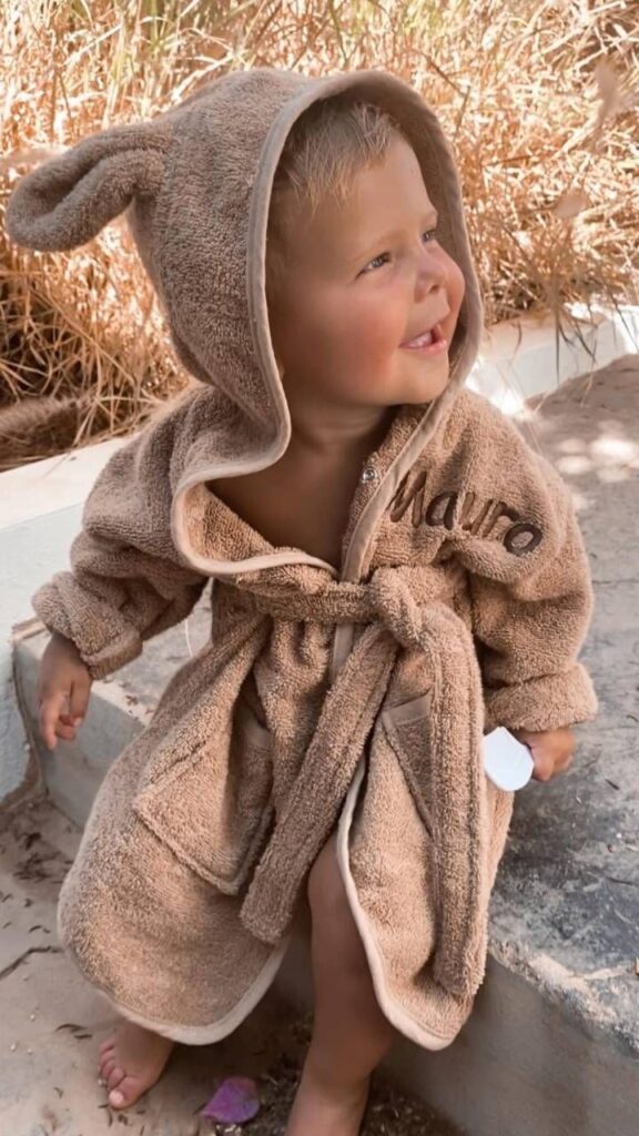 Lachend jongetje in bruine badjas met konijnenoren en geborduurd naam Mauro op zonnige stranddag